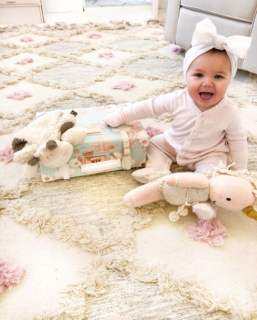 Smiling baby girl in pink pajamas with a white bow headband sitting on a soft, textured cream and pink tufted nursery rug with plush toys and a vintage-style suitcase – perfect cozy kids room decor by Rich Class Decor.