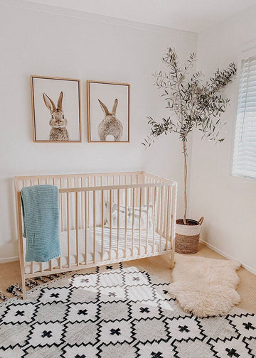 Minimalist nursery with a natural wood crib, boho-patterned rug, framed bunny artwork, and a potted olive tree, creating a cozy and modern baby room.