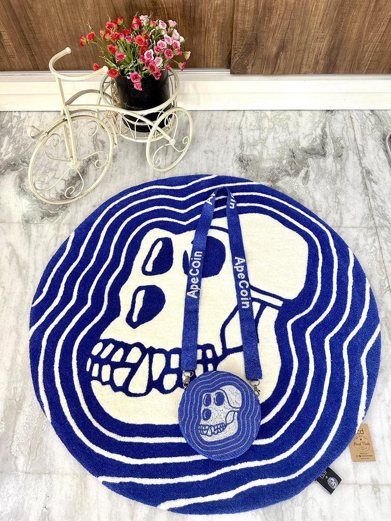 Round blue and white Apecoin-themed wool rug with matching bejewelled crossbody purse placed on top, featuring bold skull design and embroidered “Apecoin” strap, displayed on marble flooring with a decorative flower tricycle