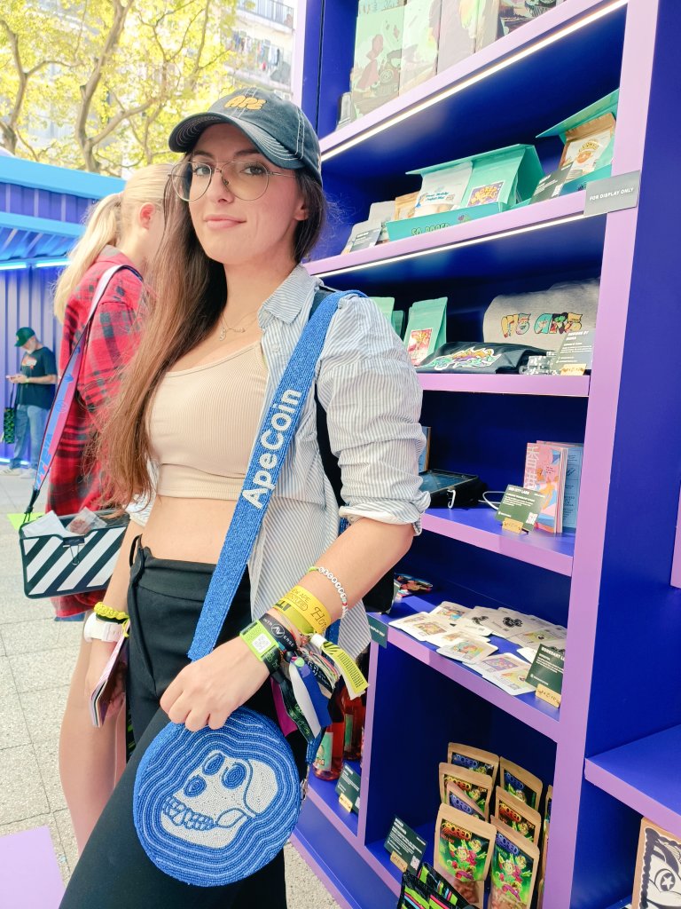 Young woman wearing a stylish bejewelled Apecoin crossbody purse with blue strap at an NFT-themed pop-up shop, standing beside vibrant purple display shelves filled with collectible merchandise.
