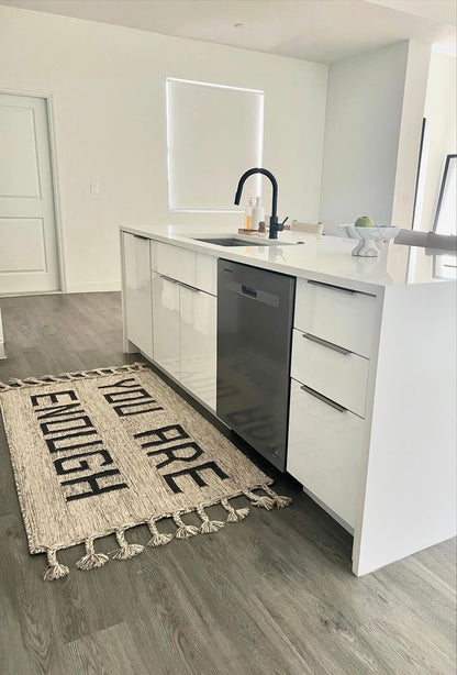A beautiful unique hand made rug is laying flat in a modern white kitchen with natural light saying You are Enough with hand made braids and neautral color making the kitchen and space bespoke while inspiring too.