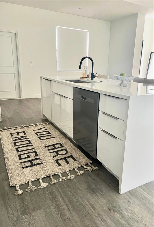 A beautiful unique hand made rug is laying flat in a modern white kitchen with natural light saying You are Enough with hand made braids and neautral color making the kitchen and space bespoke while inspiring too.