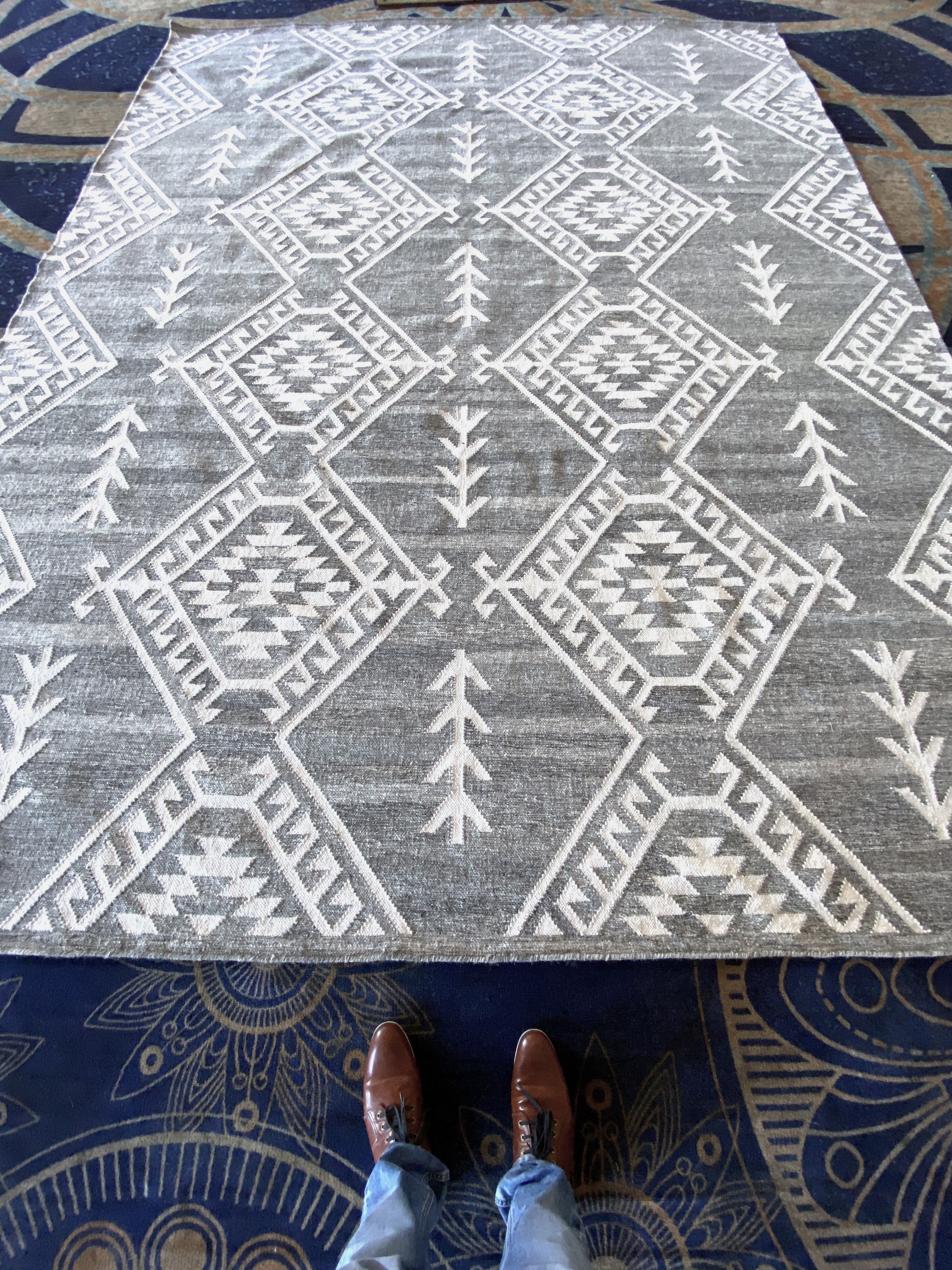 Luxury handwoven Moroccan-style area rug with a bold geometric tribal design in gray and white. A person wearing brown leather shoes and blue jeans stands at the edge, showcasing the rug’s scale and intricate craftsmanship. Perfect for contemporary, bohemian, or rustic home décor, this high-quality wool rug adds texture and warmth to any space.