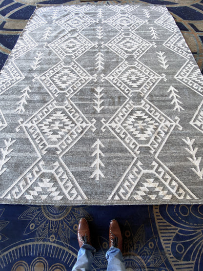 Luxury handwoven Moroccan-style area rug with a bold geometric tribal design in gray and white. A person wearing brown leather shoes and blue jeans stands at the edge, showcasing the rug’s scale and intricate craftsmanship. Perfect for contemporary, bohemian, or rustic home décor, this high-quality wool rug adds texture and warmth to any space.
