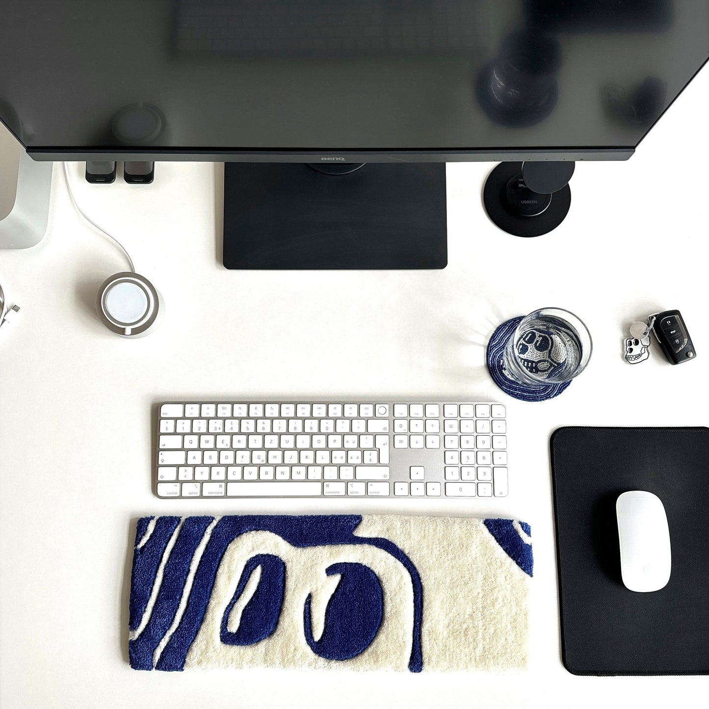 A white office table with computer keyboard glass water underneath a hand made beaded coaster in blue and white in skul shape with a custom made key board in blue and white with large eyes popping out on the side of the mouse pad looking amazing and unique 