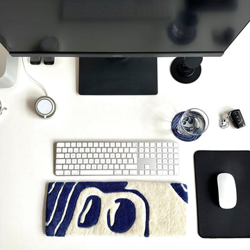 A white office table with computer keyboard glass water underneath a hand made beaded coaster in blue and white in skul shape with a custom made key board in blue and white with large eyes popping out on the side of the mouse pad looking amazing and unique 