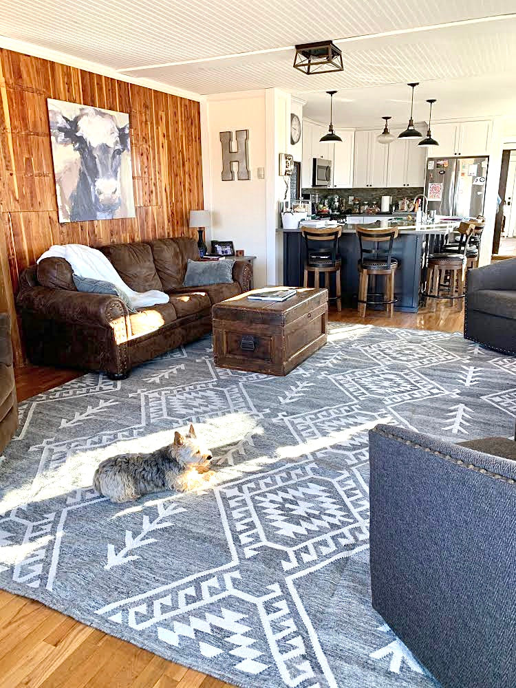 Handwoven Southwestern area rug in a modern farmhouse living room – large gray and white geometric patterned rug complementing rustic decor with a brown leather couch, wooden trunk, and open-concept kitchen. Perfect for boho, tribal, and contemporary interior design styles. High-quality, durable, and stylish home decor rug.