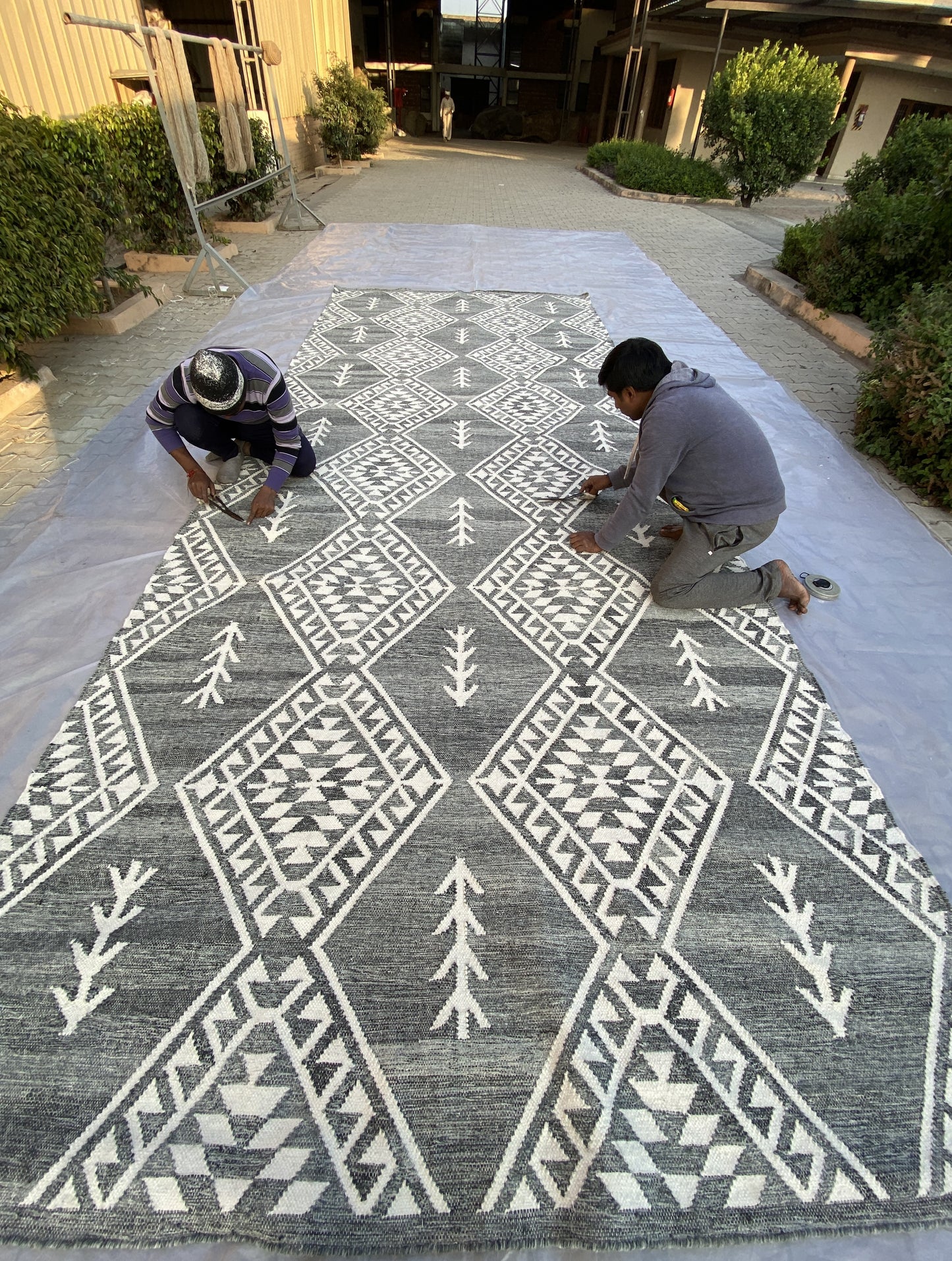 Two artisans hand-finishing a large, handcrafted black and white geometric-patterned rug outdoors, showcasing traditional craftsmanship and intricate detailing