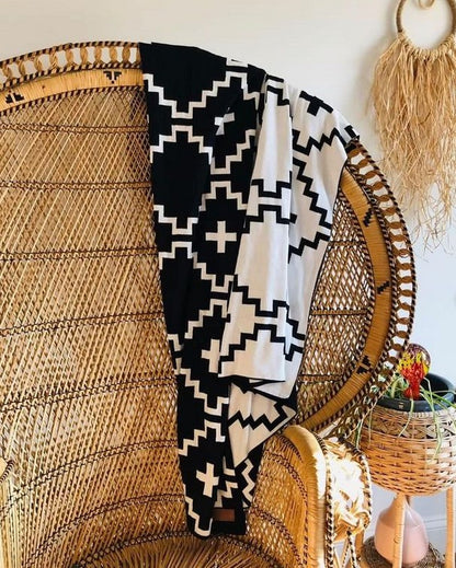 Faith Organic Cotton Throw Blanket draped over a bohemian-style wicker chair. The black and white geometric design adds a bold, modern touch to natural, earthy décor. Perfect for cozying up in a stylish home setting. Free matching pillow included for a limited time.