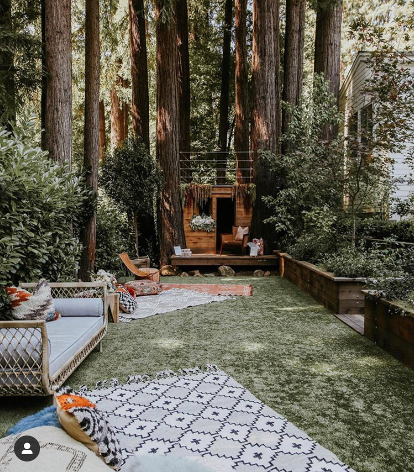 Bohemian-style outdoor lounge with woven rattan sofa, patterned rugs, and cozy cushions, set in a lush forest retreat with a wooden treehouse.