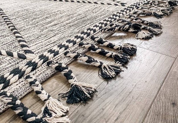 Close-up of a bohemian-style woven area rug with black and beige braided tassels on a hardwood floor, showcasing intricate craftsmanship and texture.